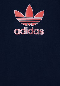 Jūras zila kokvilnas t-krekls ar izteiktu sarkanu un baltu Adidas logotipu, kas ietver trīs svītras un lapas dizainu. Gluda tekstūra.