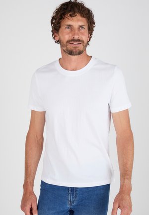 Uomo con capelli ricci castani e barba, indossa una t-shirt bianca semplice a maniche corte e jeans blu, guarda leggermente di lato.