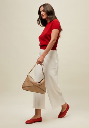 Žena v červenom tričku s krátkym rukávom, bielych širokých nohaviciach a červených balerínkach, drží béžovú kabelku, stojí pred jednoliatym béžovým pozadím.