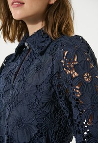 Marineblaue Spitzenbluse mit floralen Ausschneidungen, ausgestattet mit einem kragenförmigen Ausschnitt und sichtbaren Knopfakzenten auf der Vorderseite. Strukturiertes Material.