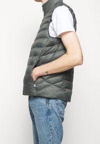 Grå, vadderad, ärmlös väst med hög krage, sidofickor och dragkedja. Buren över en vit skjorta. Texturerat, lätt material.