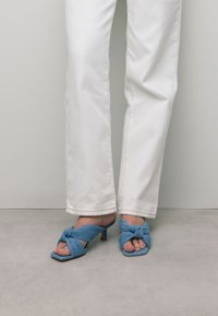 Blauwe stoffen open-teensandalen met een geknoopt ontwerp, met een kleine hak. Gecombineerd met witte rechte jeans, tegen een neutrale achtergrond.