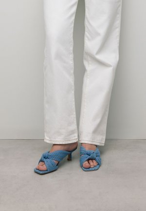 Blue fabric open-toe mules with a knotted design, featuring a small heel. Paired with white straight-leg jeans, set against a neutral background.