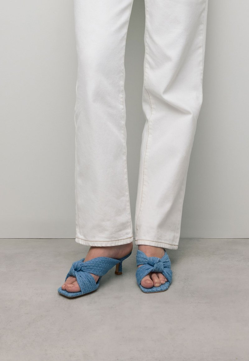 Blauwe stoffen open-teensandalen met een geknoopt ontwerp, met een kleine hak. Gecombineerd met witte rechte jeans, tegen een neutrale achtergrond.