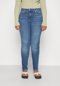 Jeans ajustados de mezclilla azul oscuro, elásticos, de talle medio, con bolsillos frontales y un ligero desgaste. Llevados con sandalias de color claro.