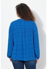 Blaue Langarmbluse mit einem Wirbelmuster, weicher Textur, angezogenem Rückendetail und lockerer Passform. Kombiniert mit dunklen Jeans.