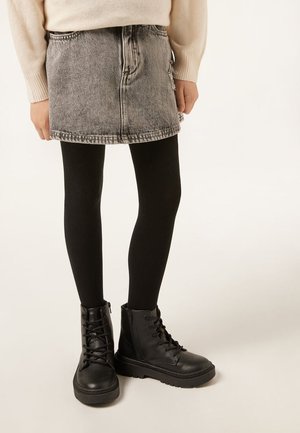 Kind trägt einen hellbeigen Pullover, einen grauen Jeansrock, schwarze Strumpfhosen und schwarze Schnürstiefeletten und steht vor einem einfarbigen Hintergrund.