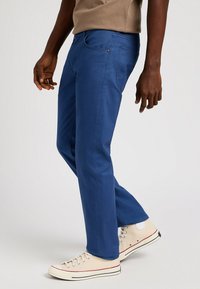 Pantaloni blu a gamba dritta in misto cotone, con tasche frontali, zip e cuciture a contrasto. Abbinati a sneakers beige alte.