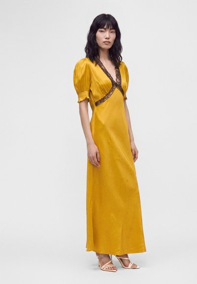 Femme portant une longue robe jaune moutarde avec des manches bouffantes et une bordure en dentelle noire, debout avec une expression neutre sur un fond clair uni.