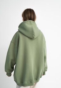 Oversized hoodie w wykonaniu z miękkiej, jasnej zielonej tkaniny. Posiada dużą kaptur, opadające ramiona oraz ściągacze na mankietach i u dołu. Gładka struktura.