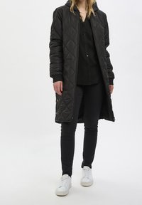 Schwarzer gesteppter langer Mantel mit Kapuze, gerippten Bündchen, zwei Seitentaschen und vollem Reißverschluss, kombiniert mit schwarzen Hosen und weißen Sneakern.