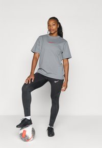 Szary koszulka sportowa krótki rękaw z czerwonym logo, w połączeniu z czarnymi legginsami o teksturowanym wzorze i marką Nike, oraz czarnymi butami sportowymi.