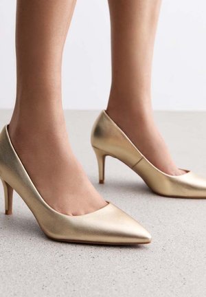 Tacones altos metálicos dorados con punta afilada y textura suave, que cuentan con un tacón delgado y un diseño minimalista.