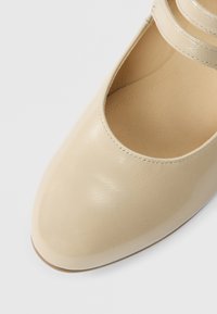 Beige laklædersko med rundet tå og to tynde ankelstropper. Glat tekstur og minimal syningsdetalje.