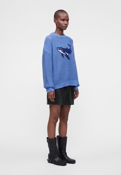 Personne portant un pull bleu tricoté avec un motif de baleine, une jupe en cuir noire et des bottes noires mi-mollet, debout devant un fond uni.