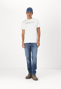 Hvid bomulds t-shirt med "TOMMY JEANS" logo, blå kasket, blå jeans og beige sko. Modellen står foran en ensfarvet hvid baggrund.