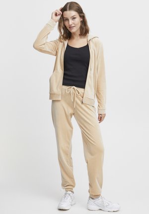 Jeune femme portant un survêtement en velours beige, un débardeur noir et des baskets blanches, se tenant avec une main touchant ses cheveux.