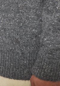Grauer gestrickter Pullover mit einer gesprenkelten Textur, der einen gerippten Saum und Bündchen aufweist. Dezente Marke auf der Brust sichtbar.