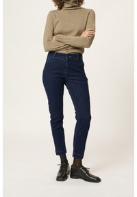 Jean en denim bleu foncé avec une coupe slim, taille haute et détails de couture minimalistes, assorti à un col roulé beige et des chaussures habillées noires.