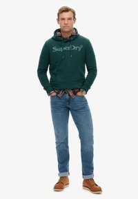 Zielona bluza z kapturem z logo, założona na czerwoną koszulę w kratę, w połączeniu z niebieskimi dżinsami i brązowymi butami sznurowanymi. Prosty design, casualowy krój.