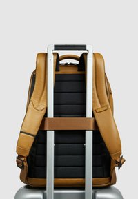 Leder-Rucksack in Hellbraun auf einem silbernen Koffer; verfügt über gepolstertes schwarzes Gewebe, verstellbare Träger und einen kontrastierenden braunen Gurtenriemen.