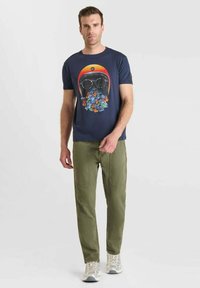 Hombre vestido con camiseta azul marino con un gráfico de casco colorido y flores, pantalones verde oliva y zapatillas gris claro, de pie frente a un fondo blanco.