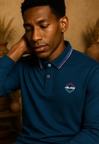 Donkerblauwe polo met lange mouwen, voorzien van een gedessineerde kraag en een logo op de linkerborst. Heeft twee knopen in contrasterende kleuren.