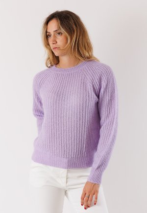 Donna con capelli mossi fino alle spalle, che indossa un maglione a coste lavanda e pantaloni bianchi, in piedi davanti a uno sfondo chiaro e semplice.