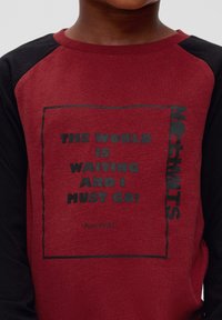 Rotes und schwarzes Langarmshirt mit einem Grafikdruck, auf dem der Text "DIE WELT WARTE UND ICH MUSS GEHEN!" in schwarzer Schrift zu sehen ist. Weicher Baumwollstoff.