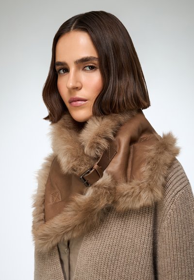 Brązowy skórzany szalik z futrzanym obszyciem, z klamrą i teksturowaną powierzchnią, noszony na beżowym dzianym ubraniu.