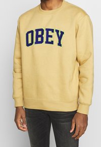 Yellow sweatshirt featuring "OBEY" in large purple and green lettering across the chest. Soft fabric, crew neckline, and ribbed cuffs.