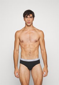 Schwarze Slips mit einem grauen Sloggi-Logo-Bund, klassischem Schnitt und glatter Textur. Das Modell steht mit Blick nach vorne vor einem einfarbigen Hintergrund.