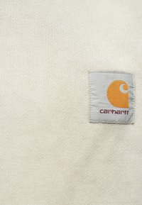 Tejido de punto beige claro que presenta un parche gris cosido con un logo naranja y texto rojo, que indica la marca "Carhartt."