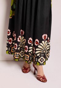 Maxi-jurk van zwarte stof met grote bloemenpatronen in roze, crème en olijfgroen, gecombineerd met rode sandaaltjes met hoge hakken.