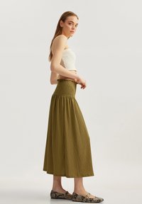 Khaki grøn, plisseret maxiskørt med smocket talje og flæsedetalje øverst, parret med en hvid cropped tanktop og mønstrede flade sko.