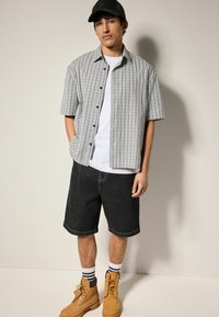 Kortærmet ternet skjorte i grøn, blå og hvid med en lomme; parret med sorte denim shorts og brune støvler, accentueret med stribede sokker.