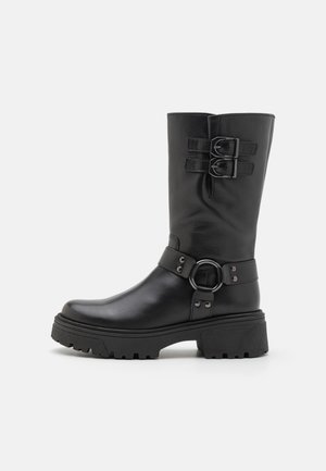 Stivale in pelle nera al ginocchio con doppie cinghie a fibbia, dettaglio di hardware circolare, suola spessa e texture liscia; presenta una punta arrotondata.
