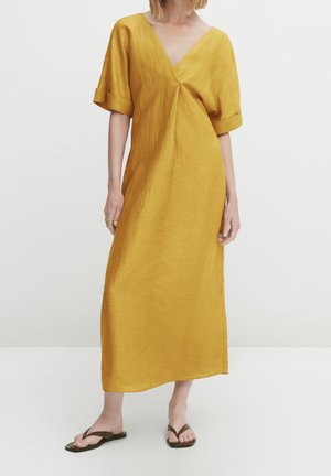 Femme portant une robe midi ample jaune moutarde à col en V, manches courtes et sandales tongs, debout sur un fond blanc.