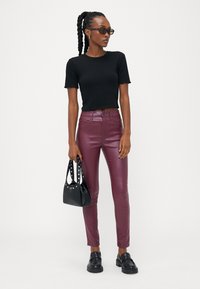 Top curto preto com nervuras e bordas em forma de ondas, combinado com calças metálicas burgundy de cintura alta. Bolsa preta com tachas e sapatos pretos robustos.