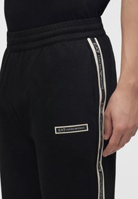 Μαύρο παντελόνι φόρμας Emporio Armani με λευκές πλευρικές ρίγες με λογότυπο και μπαλώματα κοντά στην τσέπη, φορεμένο με ταιριαστό μαύρο τοπ.