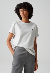 Hellgraues T-Shirt mit kurzen Ärmeln aus weichem Stoff, Rundhalsausschnitt und einem kleinen blauen Herzlogo auf der linken Brust, kombiniert mit grauen Cordhosen.