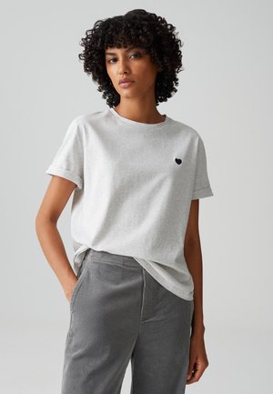 Hellgraues T-Shirt mit kurzen Ärmeln aus weichem Stoff, Rundhalsausschnitt und einem kleinen blauen Herzlogo auf der linken Brust, kombiniert mit grauen Cordhosen.