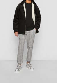 Person iklädd en svart jacka med sherpa-foder, svart skjorta, grå rutiga byxor och silvriga sneakers står mot en enkel ljusgrå bakgrund.