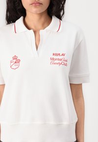 Polo blanc en tissu texturé à manches courtes, avec des accents rouges sur le col et des logos brodés sur la poitrine.