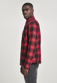 Rode en zwarte geruite flanellen shirt met lange mouwen, button-down kraag, voorzak en gemêleerde stof. Model poseert in profiel.