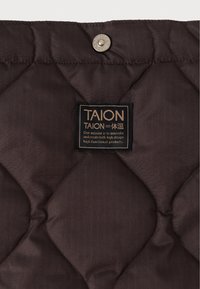 Καπιτονέ καφέ ύφασμα με μια λεπτή ριχτό υφή, με μαύρη ετικέτα με χρυσά γράμματα και ασημένιο κουμπί σνάπ στην κορυφή.