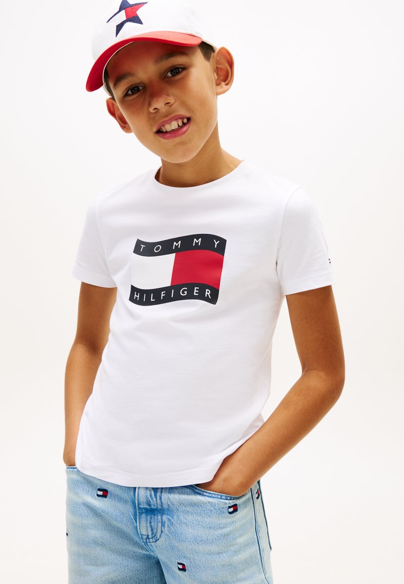 Αγόρι που φοράει λευκό μπλουζάκι Tommy Hilfiger, ανοιχτό μπλε τζιν με αυτοκόλλητα λογότυπων, και λευκό καπέλο με κόκκινο γείσο και μπλε λογότυπο αστέρι.