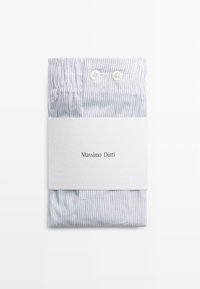 Camicia piegata con strisce blu. Un'etichetta bianca avvolge il centro, presentando il marchio. Due piccoli bottoni visibili nella parte superiore.