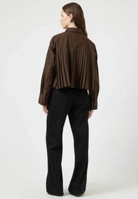 Camicia marrone plissettata con design corto e polsini con bottoni, indossata con pantaloni neri svasati. Tessuto dalla texture liscia, stile minimalista.