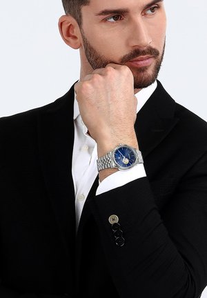 Silberne Uhr mit blauem Ziffernblatt, Edelstahlarmband und runder Gehäuseform. Die Uhr verfügt über ein Skelettziffernblatt und eine Datumsanzeige.